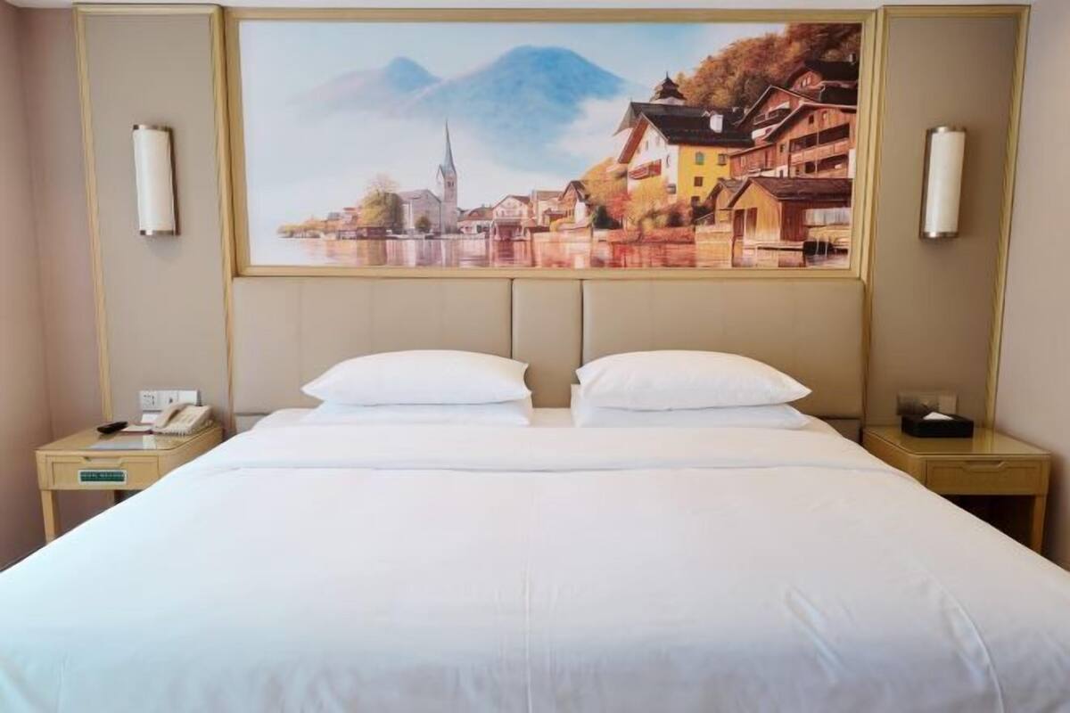 Ein Hotelzimmer mit einem großen Bett, zwei Nachttischen, einem Gemälde einer Stadtansicht und einem Telefon auf einem der Tische.