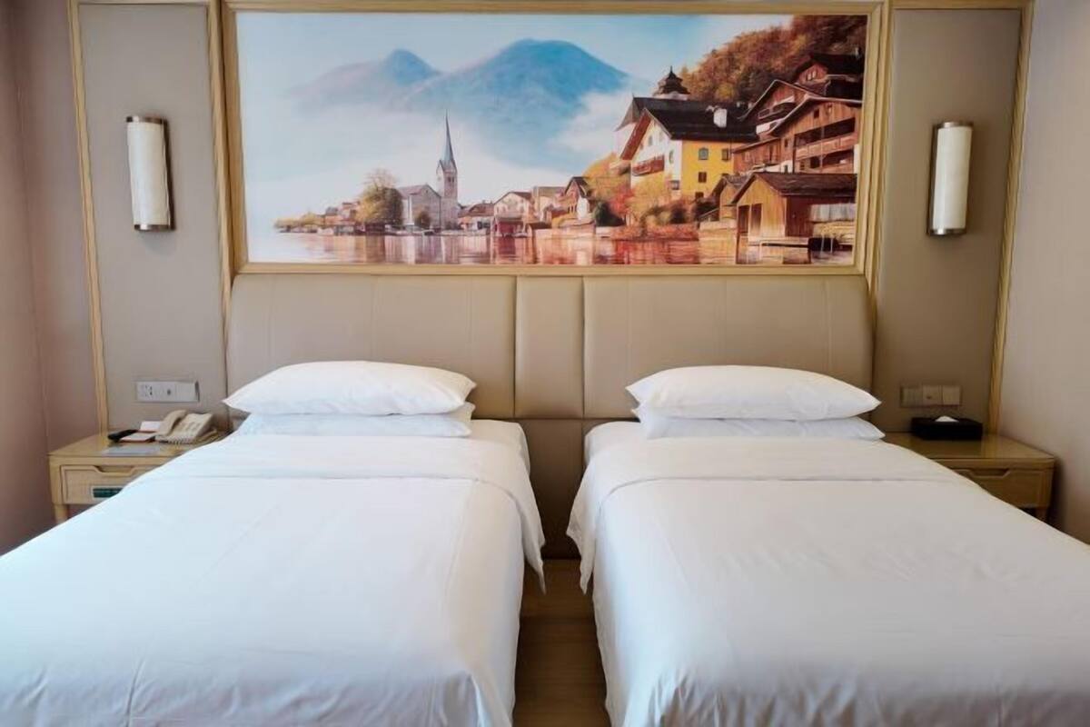 A hotel room with two beds, a painting of a town on the wall, and bedside lamps.