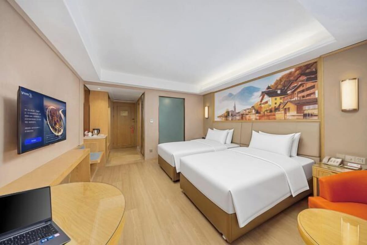 A hotel room with two beds, a desk with a laptop, a TV, and a painting on the wall.