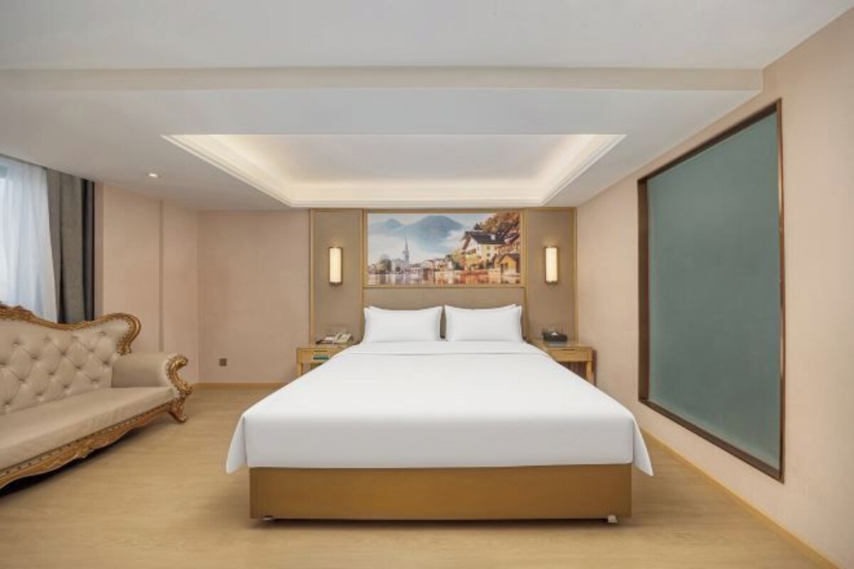 A hotel room with a large bed, a sofa, a nightstand, and a painting on the wall.