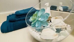 Een glazen schaal met schelpen en een blauwe bloem op een badkamermeubel.
