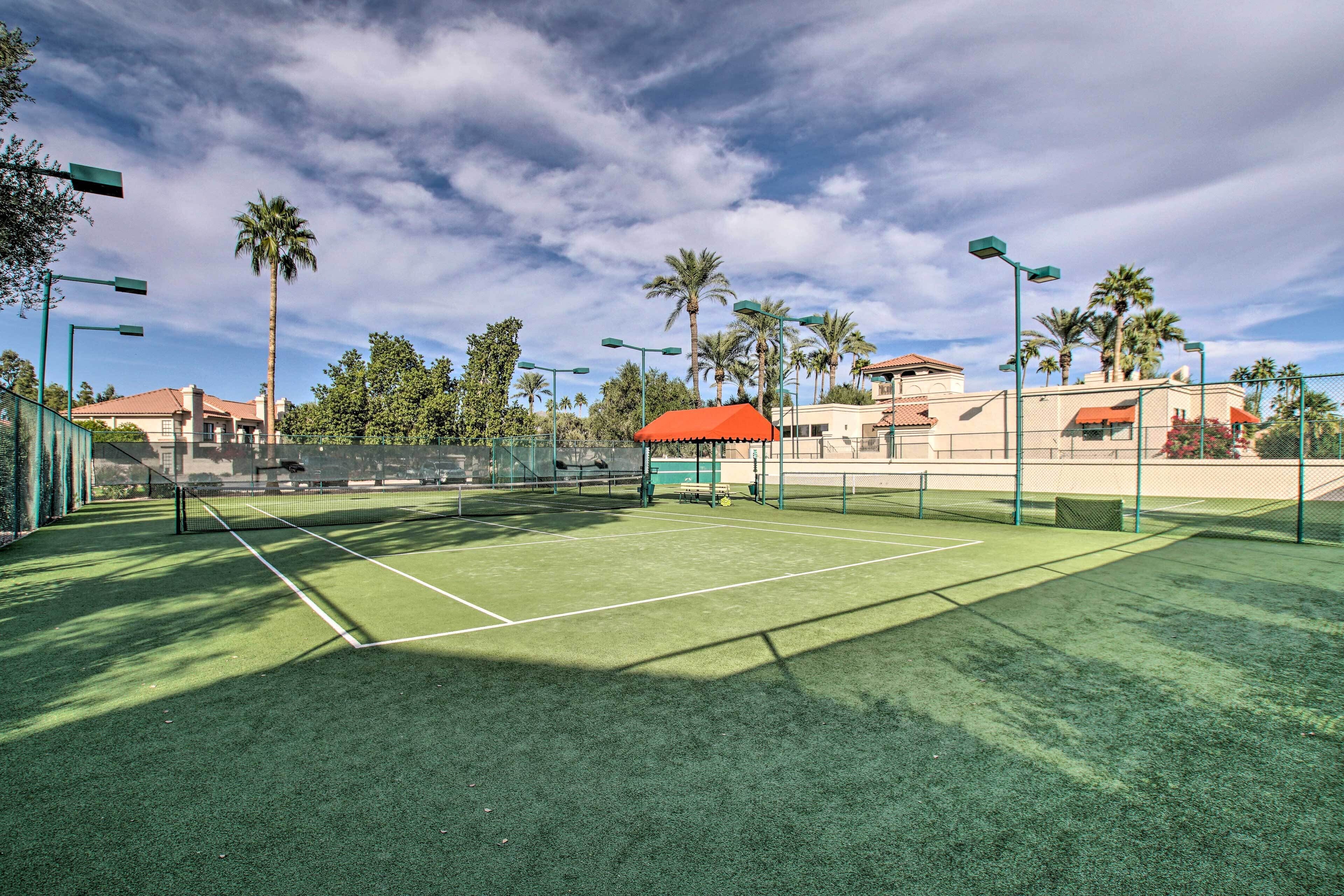 Un court de tennis en plein air, doté d’un filet, entouré de palmiers et bordé d’un bâtiment au toit rouge.