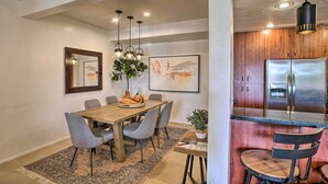 Ein Essbereich mit Holztisch, grauen Stühlen und einem gemusterten Teppich. Es gibt eine Küchenzeile mit Holzschränken und einem Edelstahlkühlschrank.