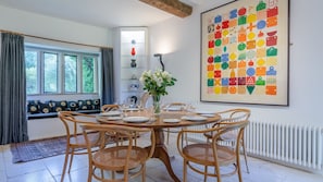 A dining area with a round wooden table set for six, surrounded by wicker chairs. There is a window with curtains, a radiator, and a large colorful artwork on the wall.