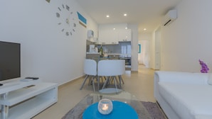 A modern living room with a TV, a glass coffee table with a candle, a white sofa, and a kitchen in the background. - Makarska Lux Apartments A4 (2+1) (Veliko Brdo, Makarska)