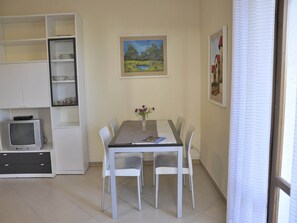 Een eethoek met een tafel, stoelen, een vaas met bloemen, een schilderij aan de muur en een televisie in een kast.