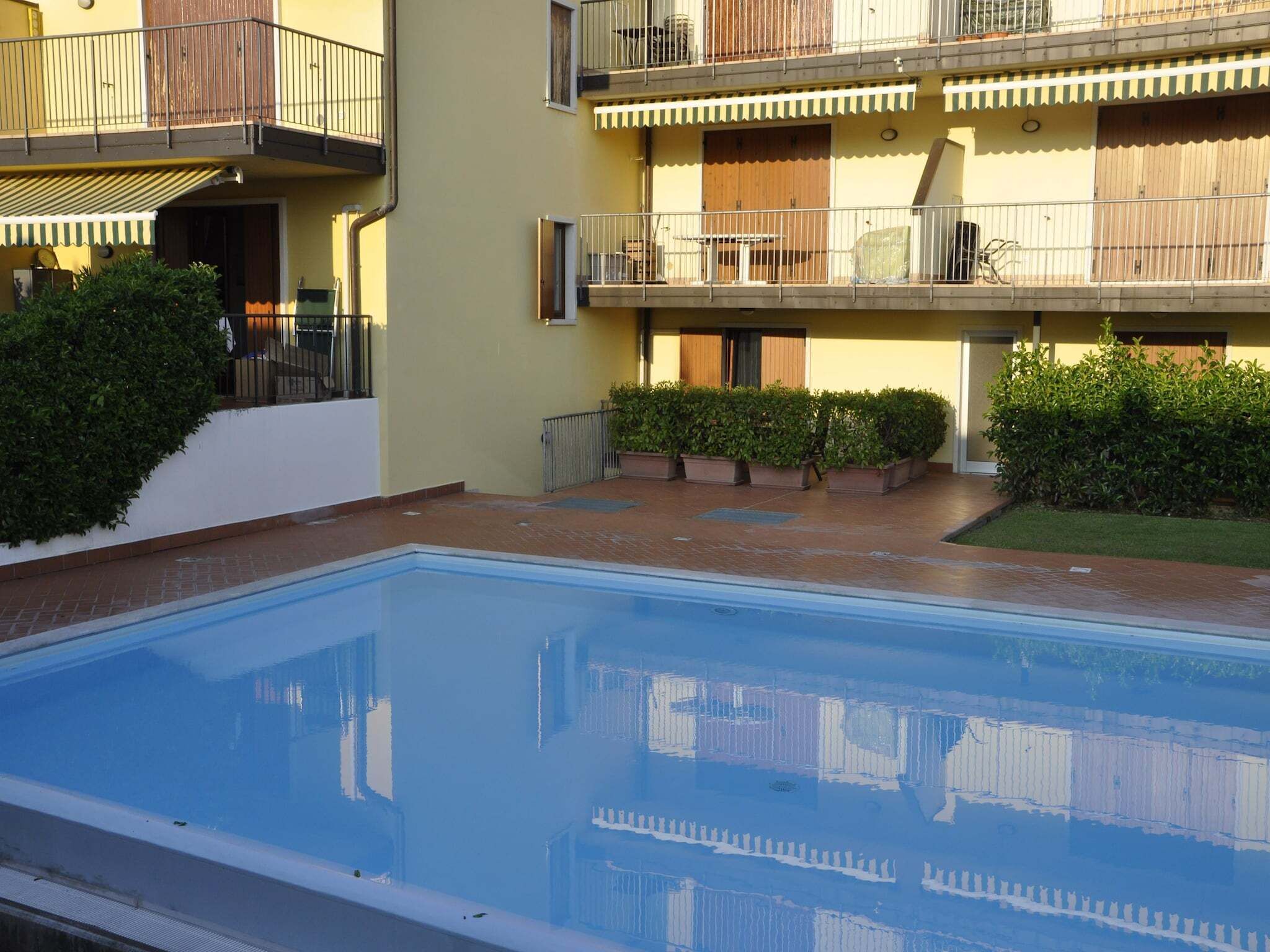 A swimming pool with a tiled deck, surrounded by potted plants and a building with balconies.