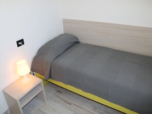 Una cama individual con cabecera de madera, una mesita de noche con lámpara y un enchufe en la pared.