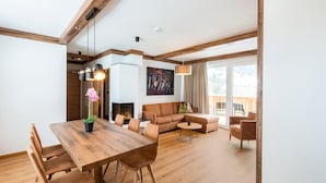 Un vaste espace de vie comprenant une table à manger en bois, un canapé et des fauteuils. On aperçoit une cheminée au fond de la pièce, et une grande fenêtre offre une vue sur l’extérieur.