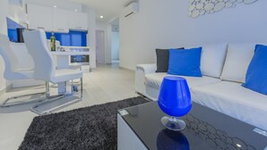 Ein modernes Wohnzimmer mit einem weißen Sofa, blauen Kissen, einem Glastisch und einem weißen Sessel.
