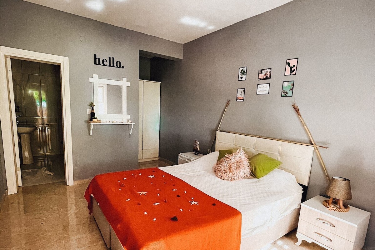 A bedroom with a bed, nightstands, a dresser, and a mirror. There is a 'hello.' sign on the wall and several framed pictures.