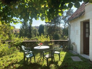 Une terrasse de jardin avec une table ronde et quatre chaises, entourée d’une végétation luxuriante et d’un mur de pierre.