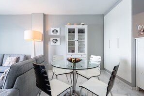 Een moderne eethoek met een glazen tafel, witte stoelen, een grijze bank en een witte kast met glazen deuren.