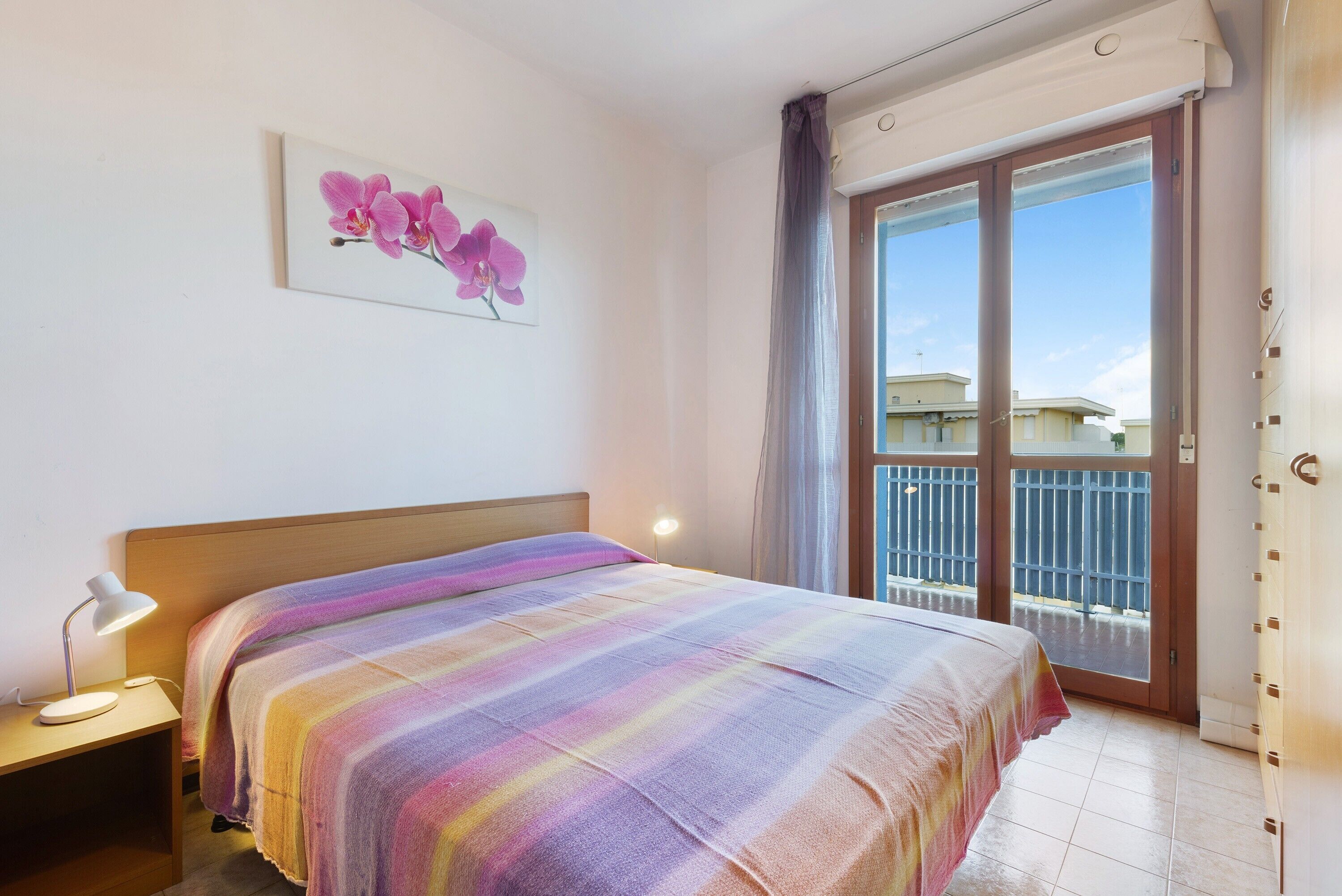 A bedroom with a bed, a nightstand, a lamp, a window with curtains, and a balcony.