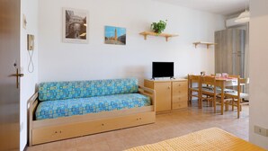 Ett litet rum med en soffa, en tv, ett matbord med stolar och en liten växt på en hylla.
