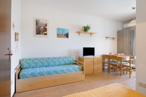 A compact room with a sofa, a TV, a dining table with chairs, and a small plant on a shelf.