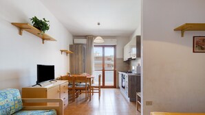 Ett kompakt bostadsutrymme med en soffa, ett matbord, en TV, ett köksutrymme och en skjutdörr i glas som leder ut till en balkong.