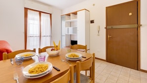 A dining area with a wooden table, chairs, and plates of food. There is a door with a handle and a shelf unit against the wall. - Studio "Valbella A" in Residence with 12 Pools (Bibione)