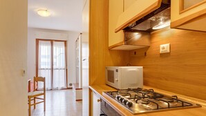 A kitchen with a gas stove, microwave, wooden cabinets, and a window with curtains. - Studio "Valbella A" in Residence with 12 Pools (Bibione)
