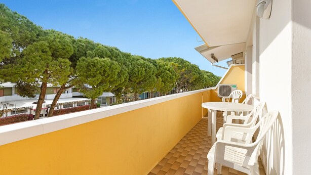 A balcony with a yellow wall, white chairs, a small table, and a view of trees and buildings. - Apartment "Condominio Simma 1B6" with A/C & Terrace (Bibione)