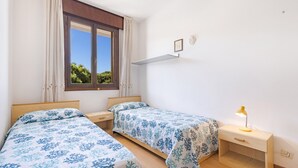 A bedroom with two beds, a window with curtains, a bedside table with a lamp, and a wall-mounted shelf.