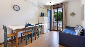 A room with a dining table, chairs, a sofa, a TV, a refrigerator, and a window with curtains.