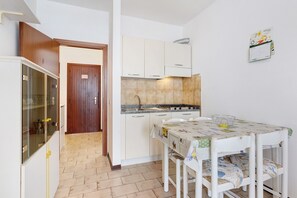 A compact kitchen with white cabinets, a wooden door, a tiled backsplash, and a dining table with a patterned tablecloth.