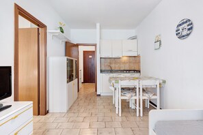 A compact kitchen with white cabinets, a dining table with chairs, a TV on a dresser, and a clock on the wall.