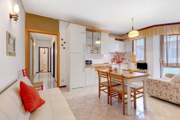 A modern kitchen with white cabinets, a dining table with chairs, a sofa with a red pillow, and a TV.