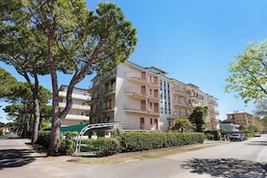 A multi-story residential building with balconies, a green hedge, and parked cars.
