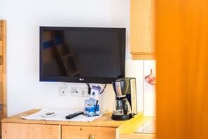 Un televisor, una cafetera y un control remoto sobre un escritorio de madera, con una pared blanca de fondo.