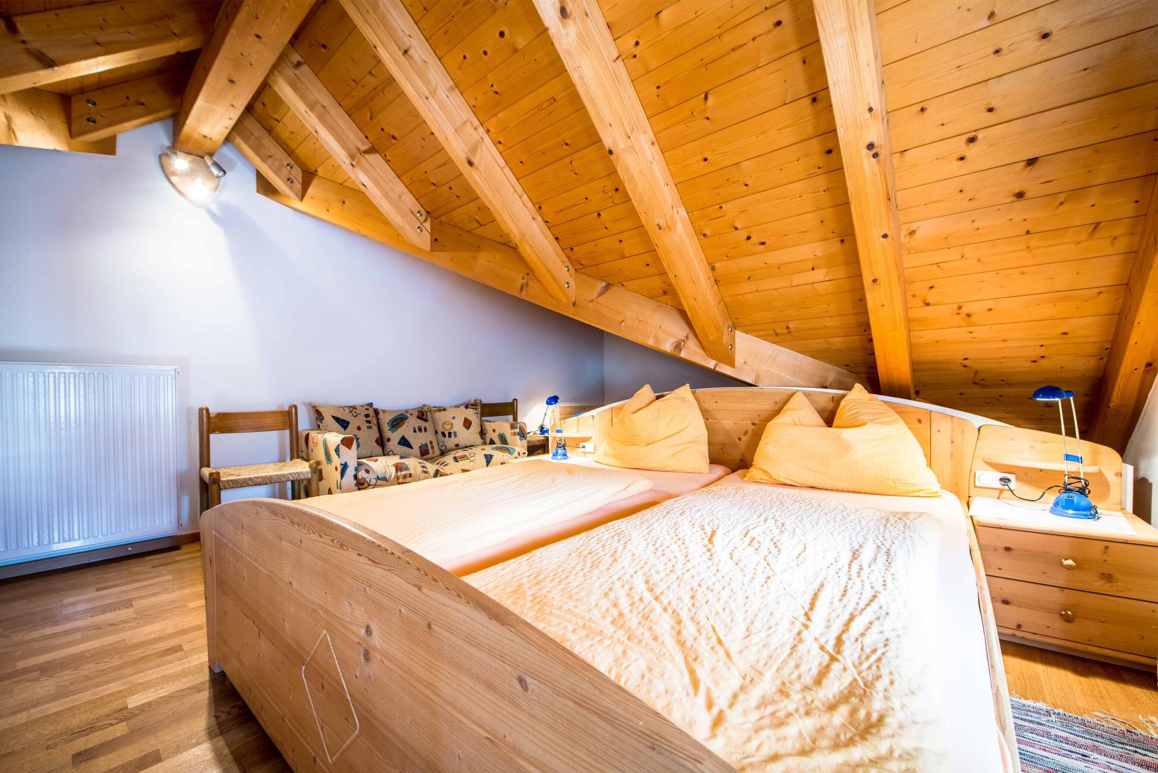 A wooden ceiling with a sloped design, a bed with a patterned bedspread, a nightstand with a lamp, and a chair with a patterned cushion.
