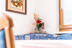Ein Zimmer mit einem Tisch, der mit einem blauen Tuch bedeckt ist, einer Blumenvase, einem gerahmten Bild an der Wand und einem Fenster mit Spitzenvorhängen.
