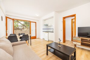 A modern living room with a sofa, dining table, and chairs, a kitchen with white cabinets, and a TV on a wooden stand.
