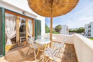 Un balcon avec une table et des chaises, un parasol en chaume et une vue sur les bâtiments environnants.