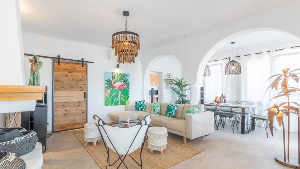 En moderne stue med sofa, spisebord, stole, en lysekrone og et stort vindue.