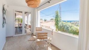 En balkon med en fletlampe, et spisebord med stole og udsigt til havet og bygninger.