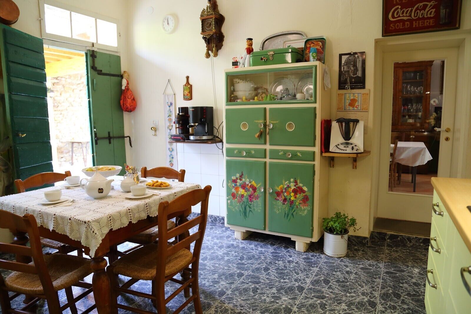 A dining area with a table set for two, a green cabinet with floral decorations, and a clock on the wall.