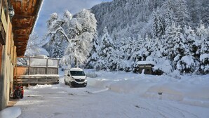Eine verschneite Bergstraße mit einem weißen Auto vor einem Holzbau.