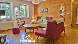 Ein gemütliches Wohnzimmer mit Holzboden, einem roten Sofa, einem grünen Sofa und einem Holztisch. Zwei Fenster mit Vorhängen, eine Heizung und ein Bild an der Wand sind ebenfalls zu sehen.
