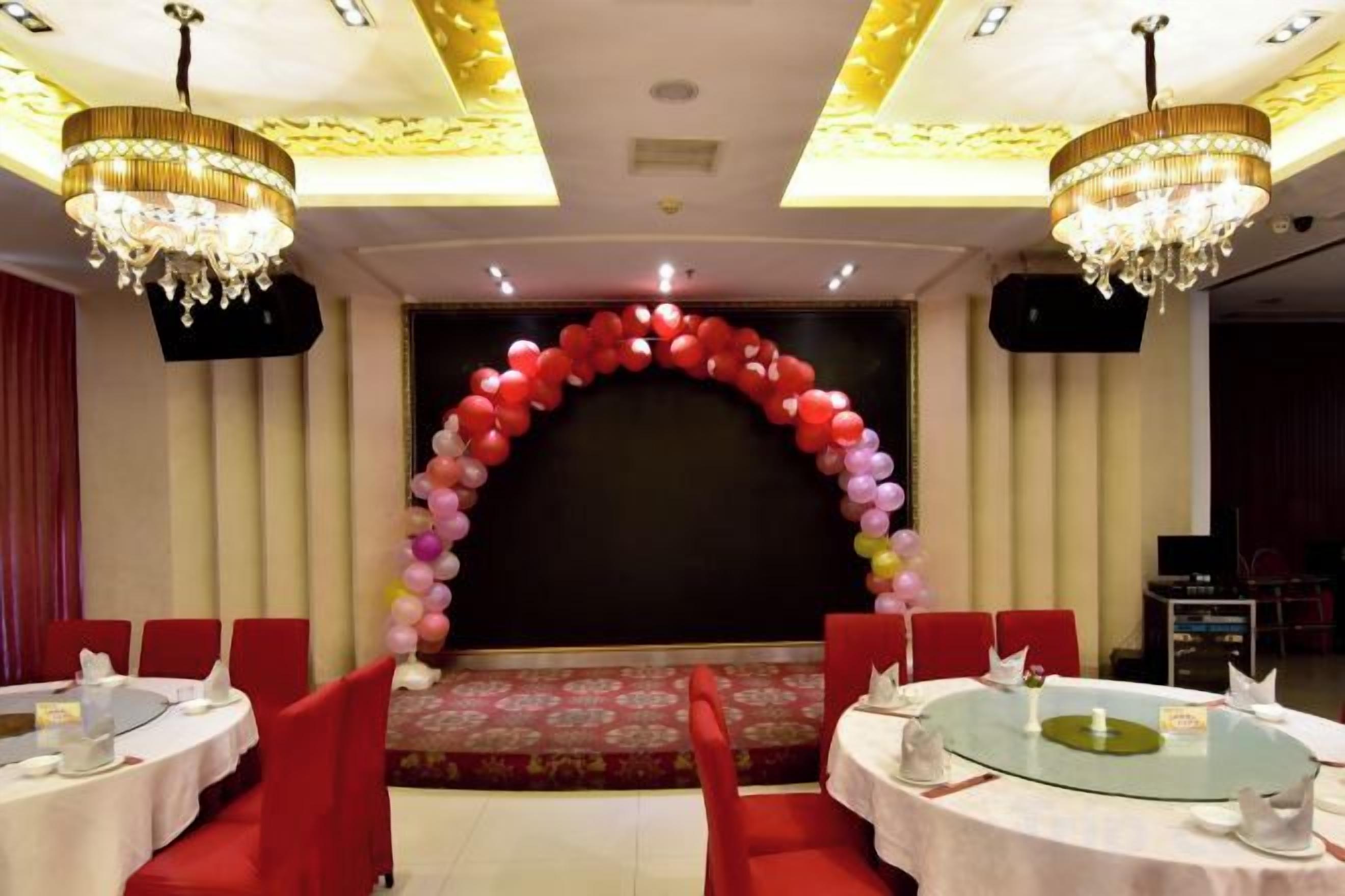 A banquet hall with round tables, red chairs, a stage with a balloon arch, chandeliers, and a black backdrop.