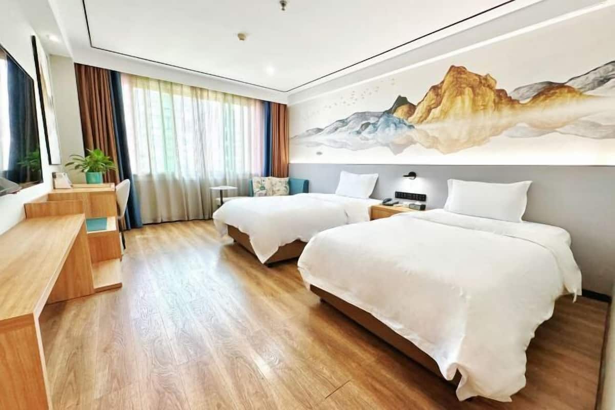 A hotel room with two beds, a desk, a chair, a TV, and a painting of mountains on the wall.