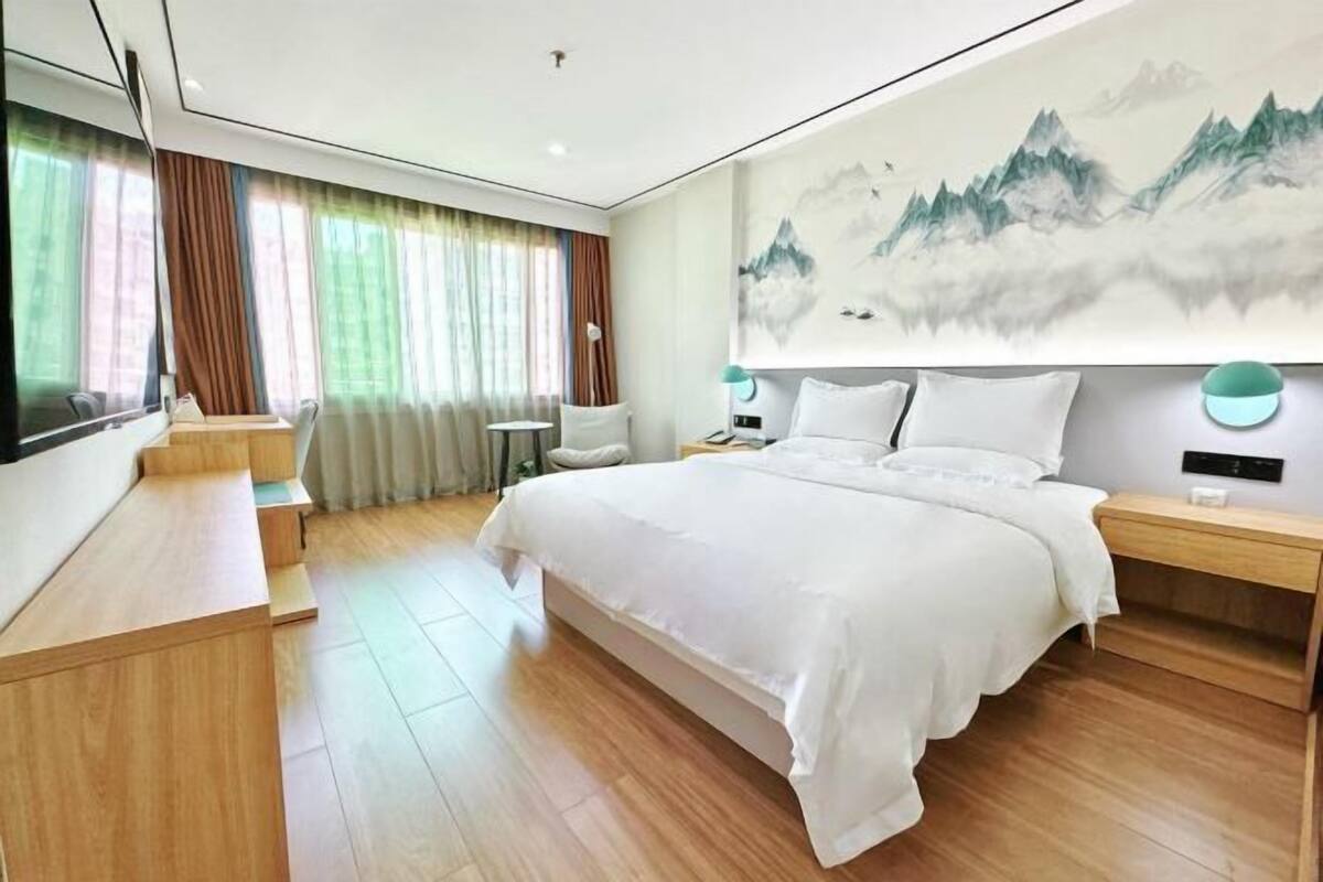 A modern hotel room with a large bed, wooden flooring, a desk, and a large painting of mountains.
