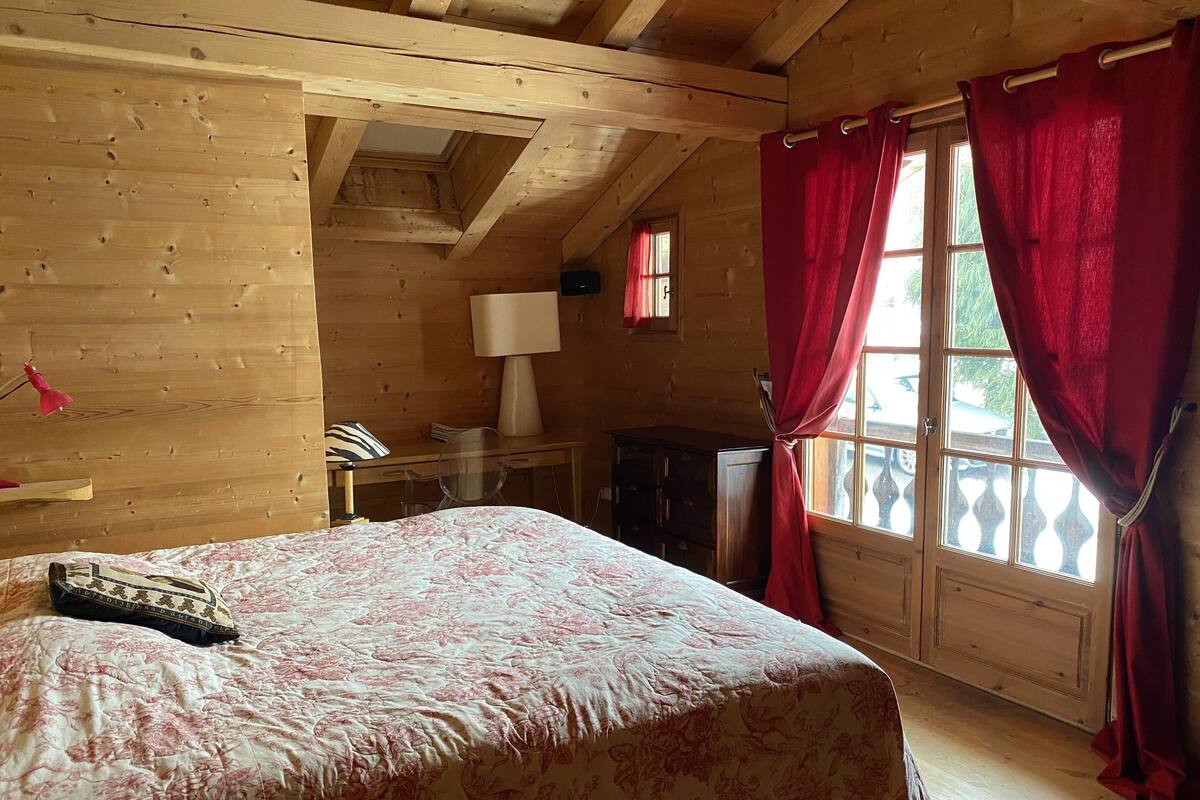 A wooden cabin room with a bed, a desk, and a window with red curtains.