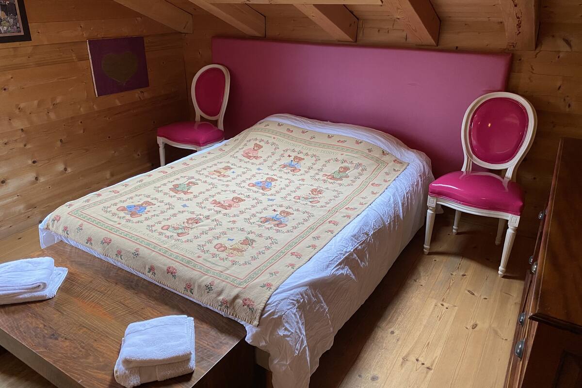 A cozy attic bedroom with a wooden ceiling, a pink upholstered headboard, two matching pink chairs, a bed with a floral quilt, and a wooden table with two folded white towels.