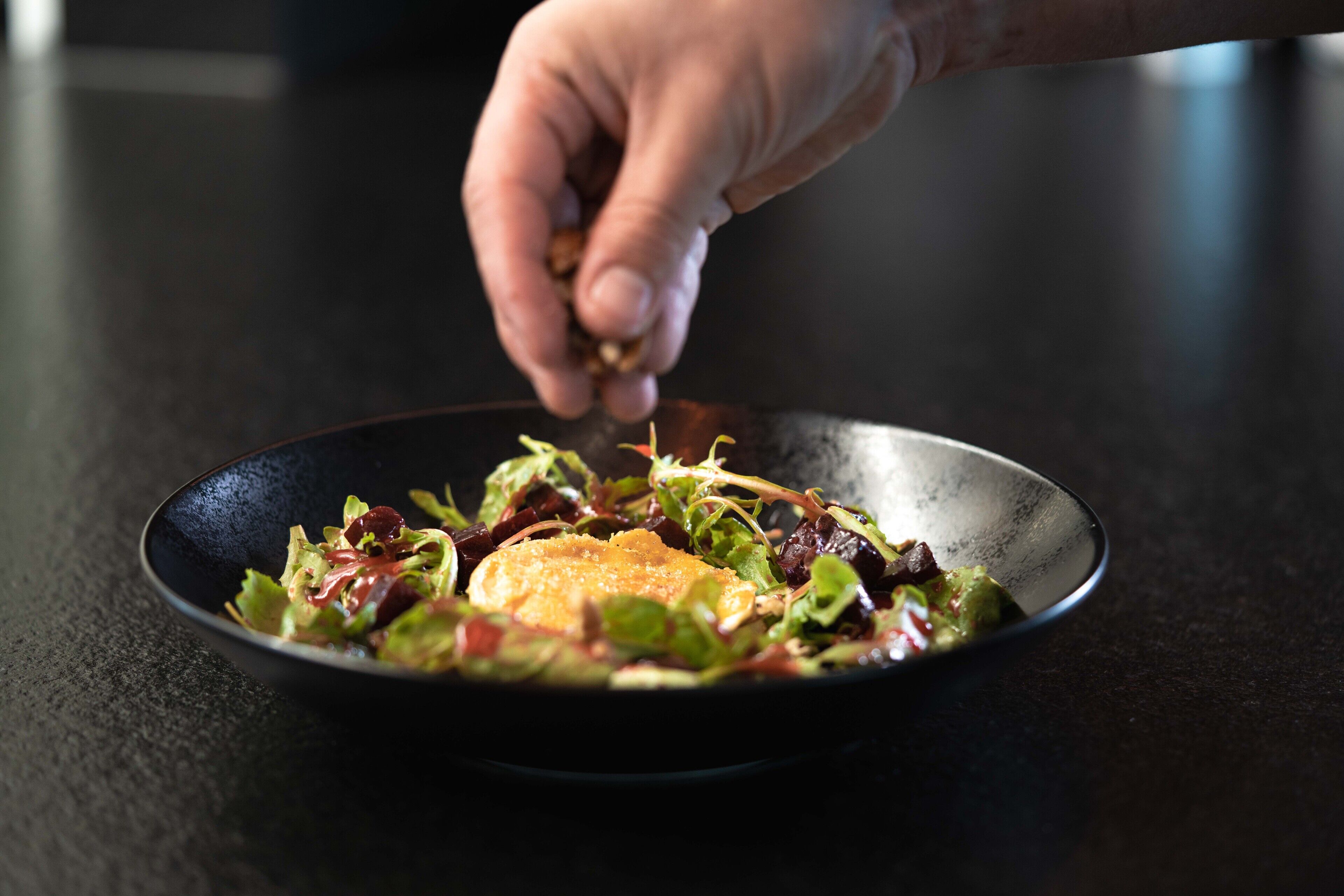 Una mano esparciendo condimentos sobre una ensalada en un tazón negro sobre una superficie oscura.