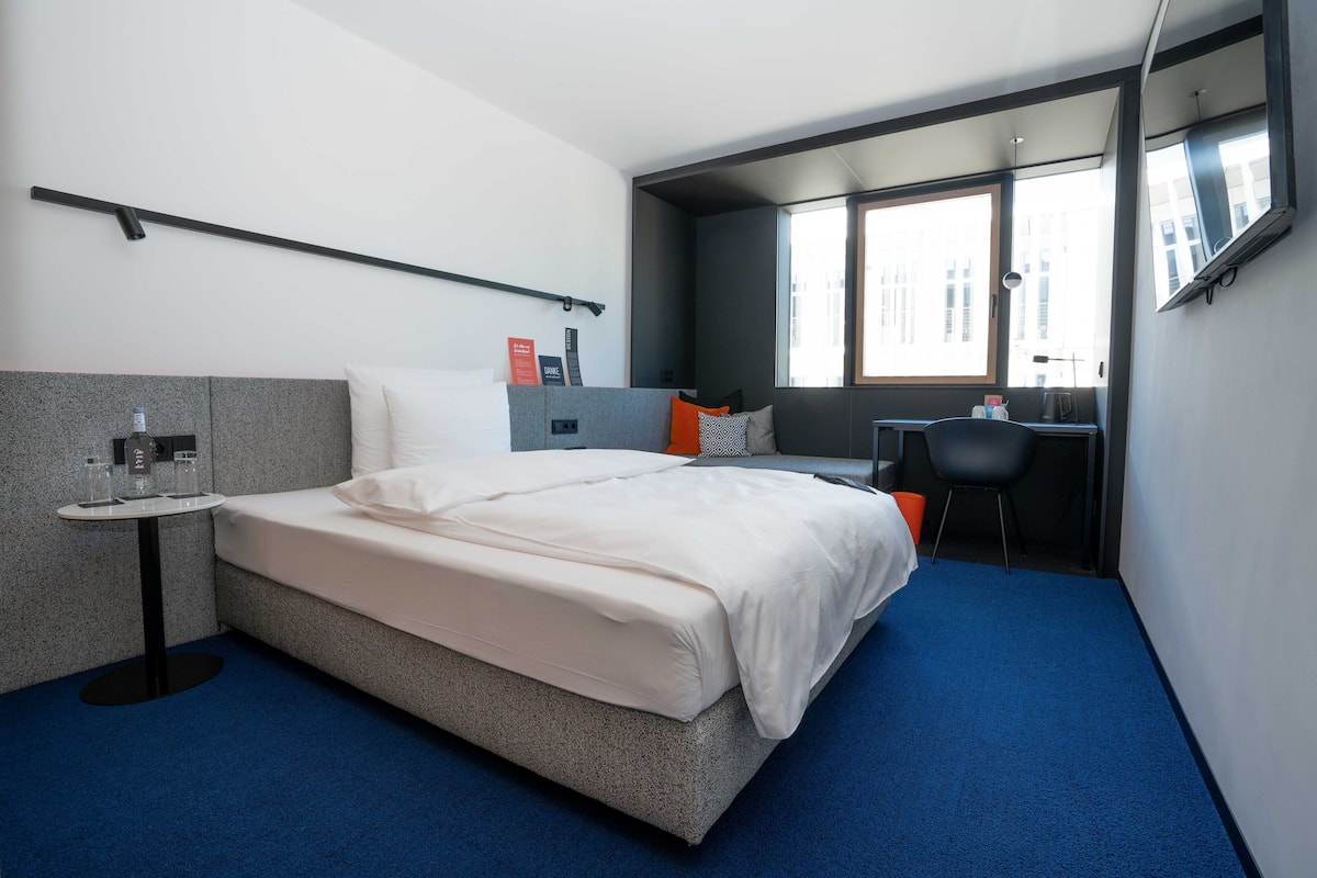 Ett modernt hotellrum med en stor säng, ett skrivbord och en stol. Det finns ett fönster som släpper in naturligt ljus och erbjuder utsikt över staden.
