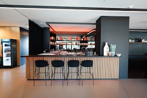 Zona de bar moderna con una barra de madera, tres taburetes negros y estantes llenos de botellas y copas.