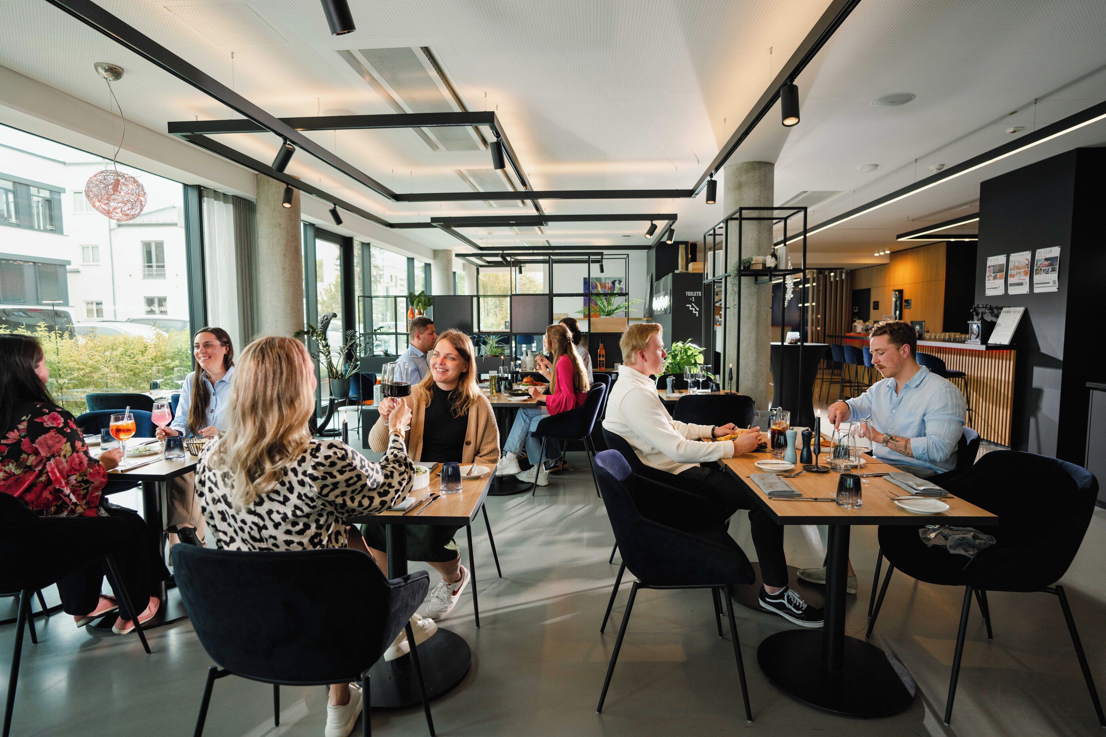 Et moderne spiseområde med borde, stole, store vinduer og indeplanter.