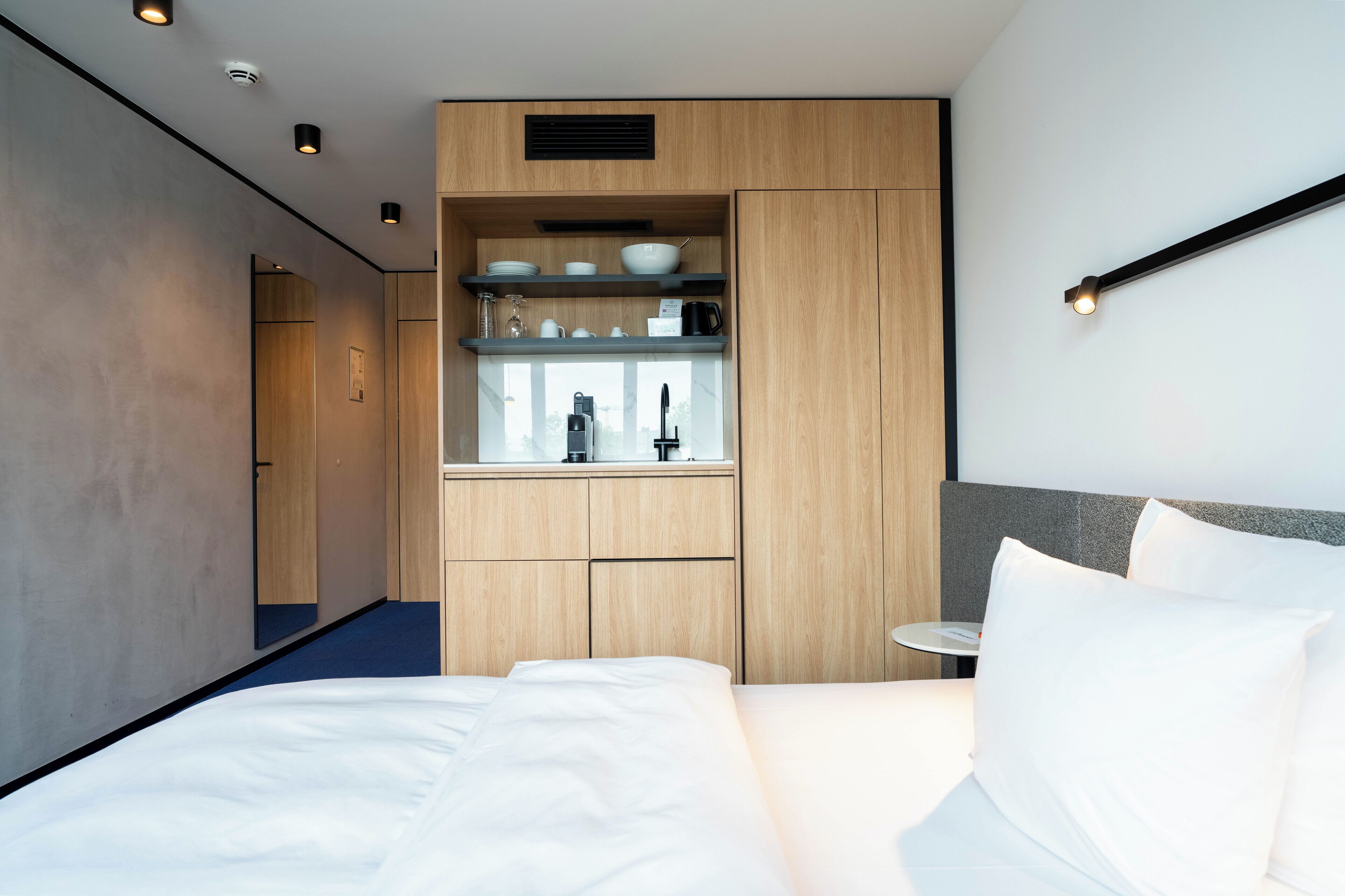 Una habitación de hotel moderna con una cama grande, una kitchenette integrada con cafetera y una pequeña mesa redonda.